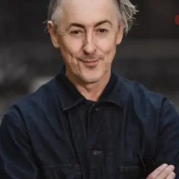 Alan Cumming stands with arms crossed, smiling slightly at the camera against a blurred dark background.