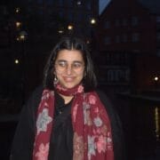 A person with short dark hair and glasses, wearing a black jacket and a red floral scarf, stands outdoors by water at dusk with buildings in the background.