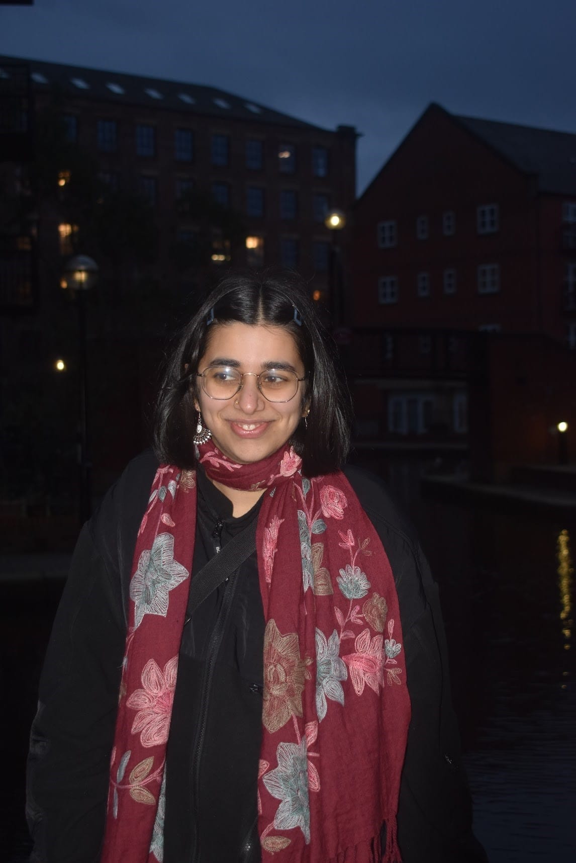 A person with short dark hair and glasses, wearing a black jacket and a red floral scarf, stands outdoors by water at dusk with buildings in the background.