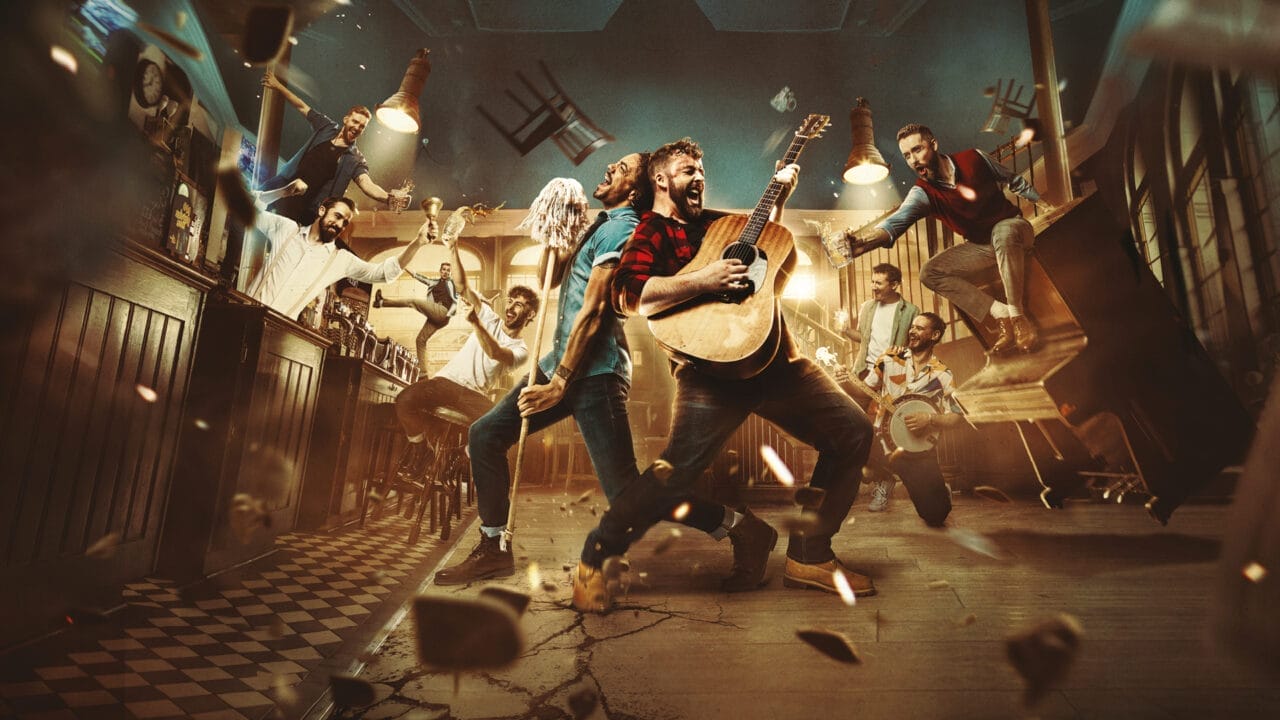 A group of people, including performers from The Choir of Man, are playing musical instruments and dancing energetically in a bar, with chairs and debris flying around, suggesting a lively and chaotic scene.