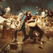 A group of people, including performers from The Choir of Man, are playing musical instruments and dancing energetically in a bar, with chairs and debris flying around, suggesting a lively and chaotic scene.