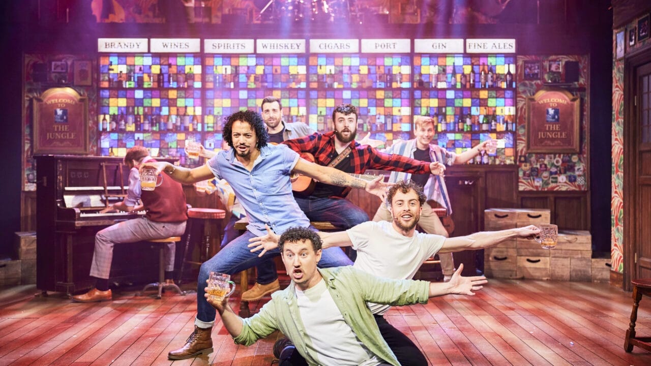 The Choir of Man performers energize the stage in a lively bar setting, holding drinks under colourful lights as the band plays in the background.