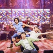 The Choir of Man performers energize the stage in a lively bar setting, holding drinks under colourful lights as the band plays in the background.