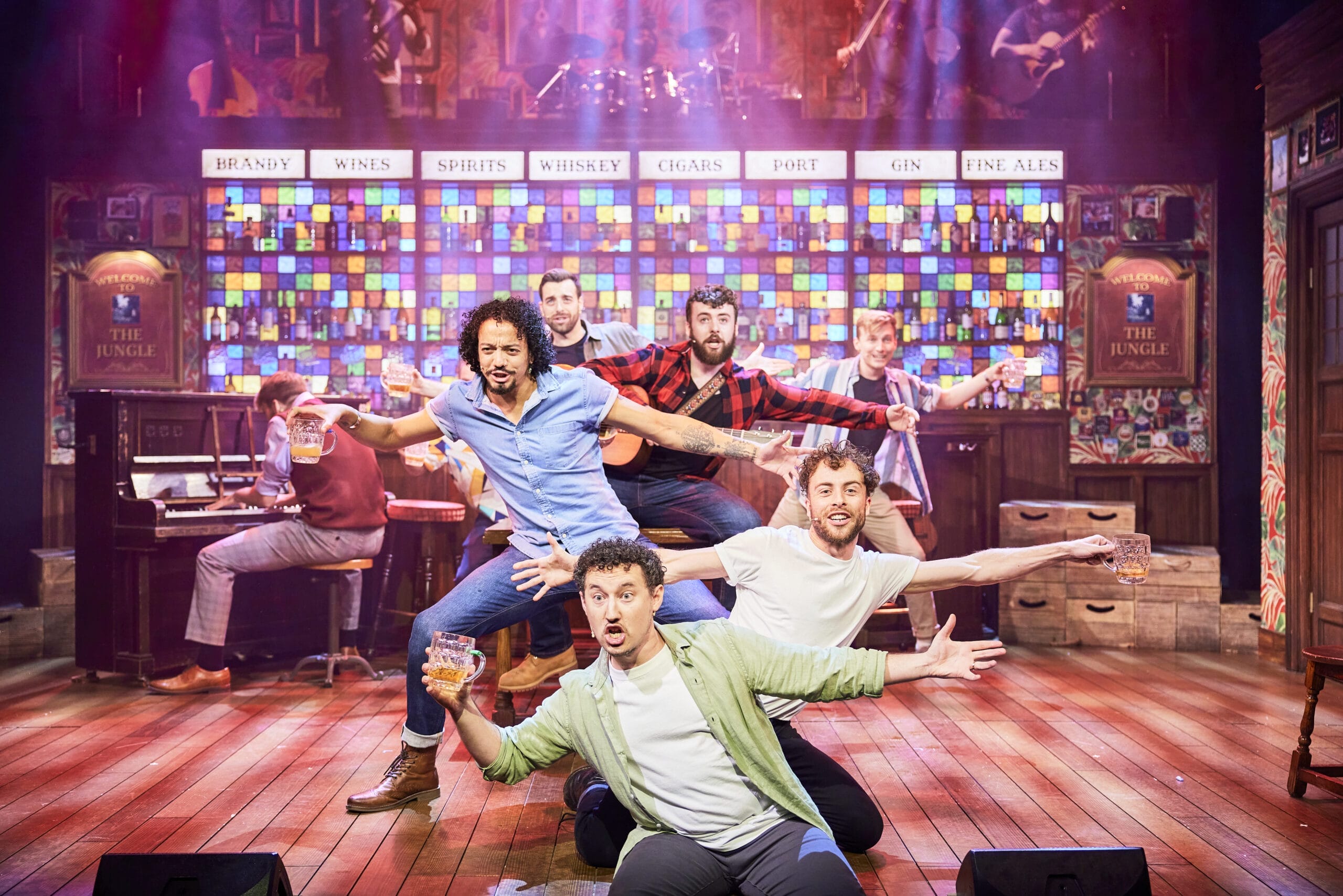 The Choir of Man performers energize the stage in a lively bar setting, holding drinks under colourful lights as the band plays in the background.