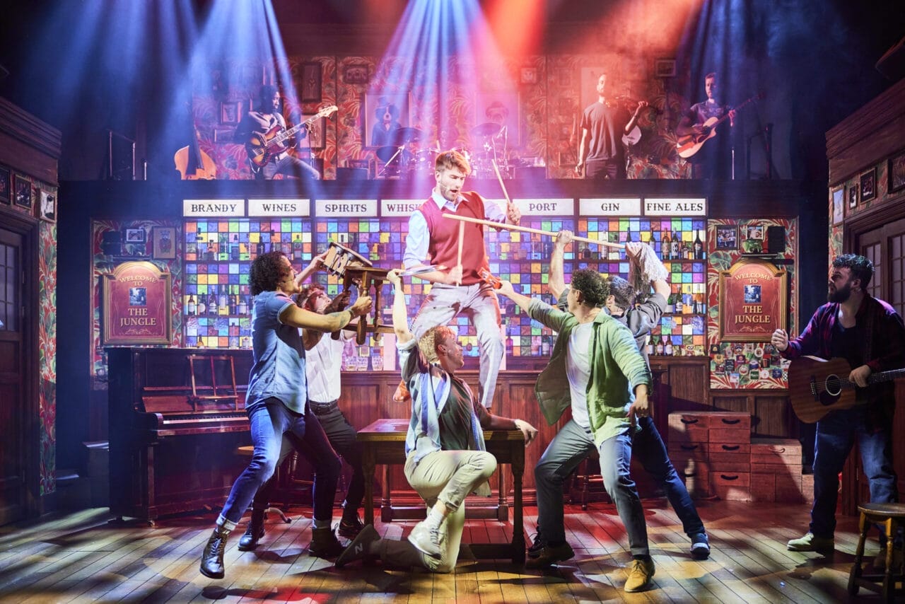 Actors from The Choir of Man perform a lively scene on stage in a pub setting, with one person elevated on a table and others holding props; musicians play in the background under colorful stage lights.