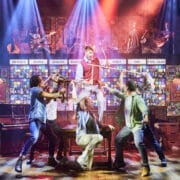 Actors from The Choir of Man perform a lively scene on stage in a pub setting, with one person elevated on a table and others holding props; musicians play in the background under colorful stage lights.