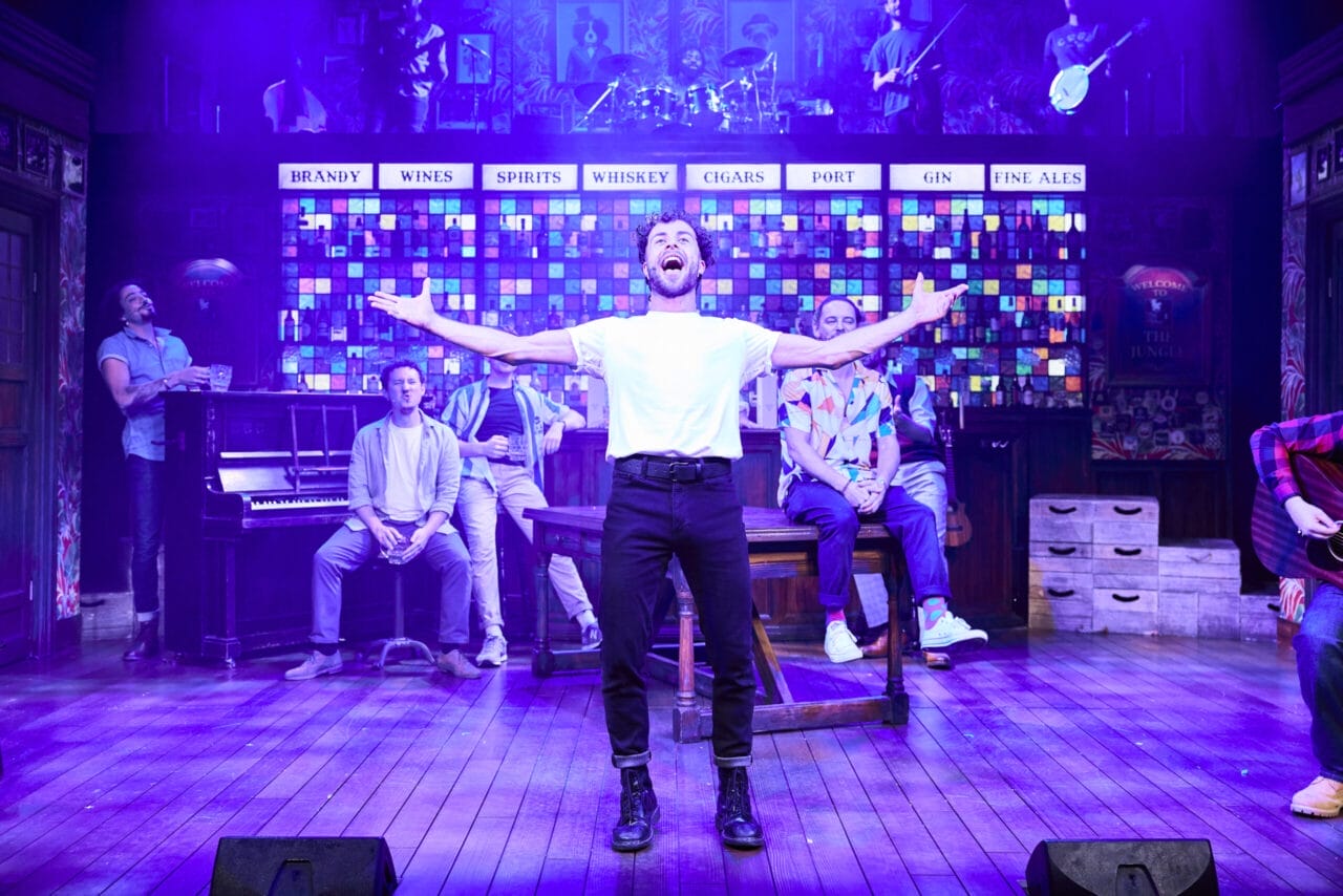 A man stands center stage with arms outstretched, surrounded by other performers from The Choir of Man in a colourful, bar-themed musical set.