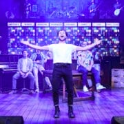 A man stands center stage with arms outstretched, surrounded by other performers from The Choir of Man in a colourful, bar-themed musical set.