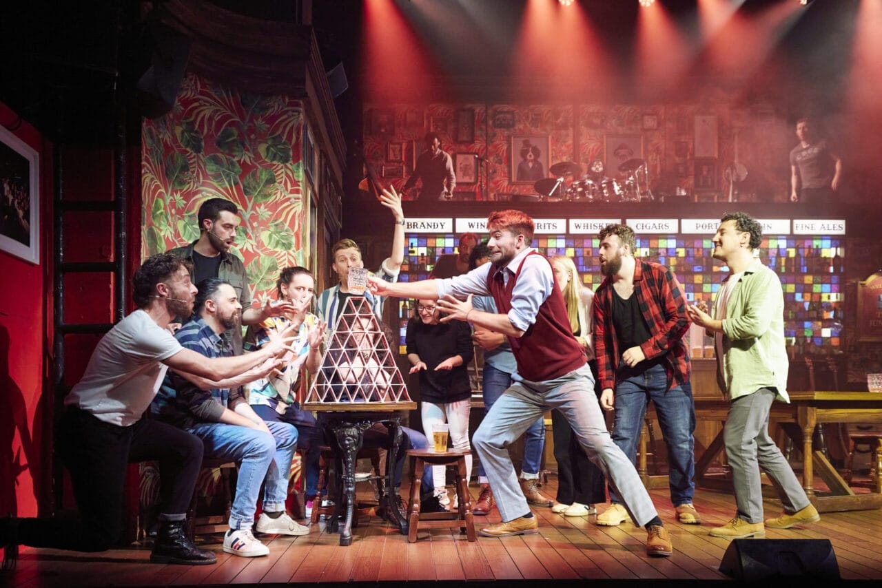 A group of people on stage dramatically reach toward a tall pyramid of stacked cards under bright stage lights, with The Choir of Man performing against a colourful bar scene backdrop.
