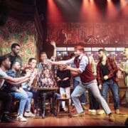 A group of people on stage dramatically reach toward a tall pyramid of stacked cards under bright stage lights, with The Choir of Man performing against a colourful bar scene backdrop.