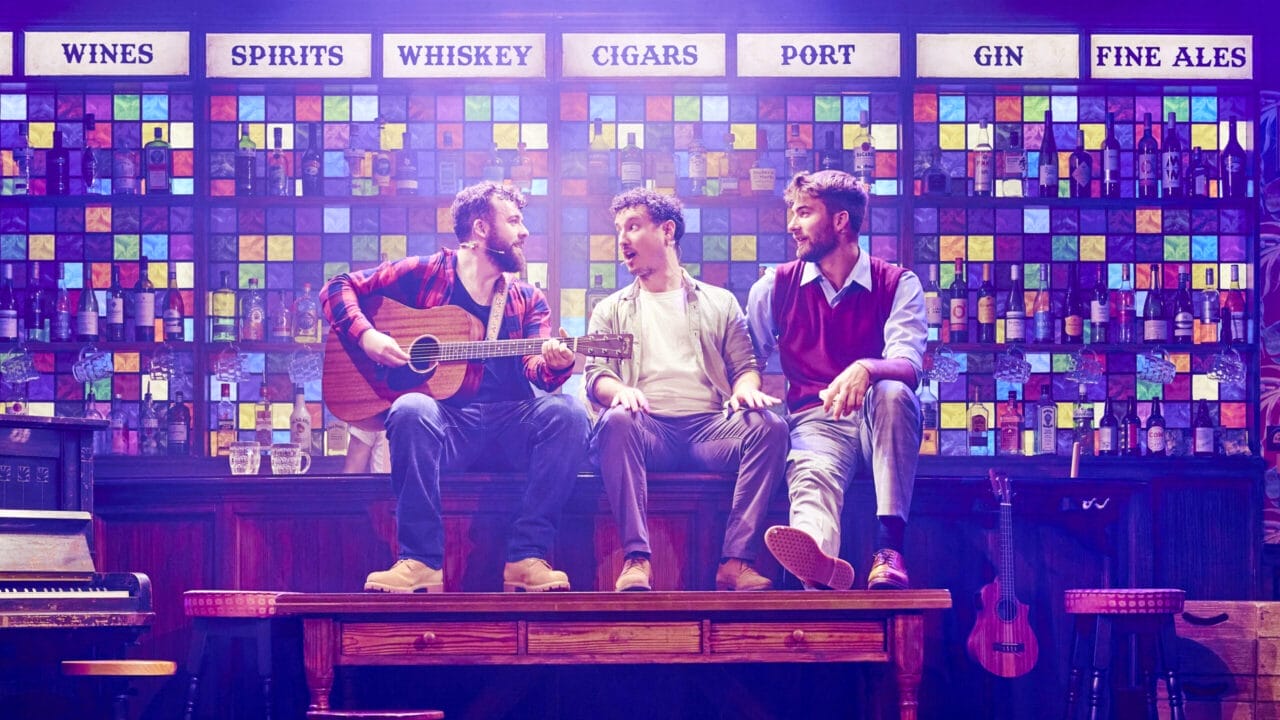 Three men sit on a wooden table in front of a colourful bar wall display; one strums a guitar while the others converse under signs for various drinks, reminiscent of The Choir of Man’s lively pub atmosphere.