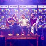 Three men sit on a wooden table in front of a colourful bar wall display; one strums a guitar while the others converse under signs for various drinks, reminiscent of The Choir of Man’s lively pub atmosphere.
