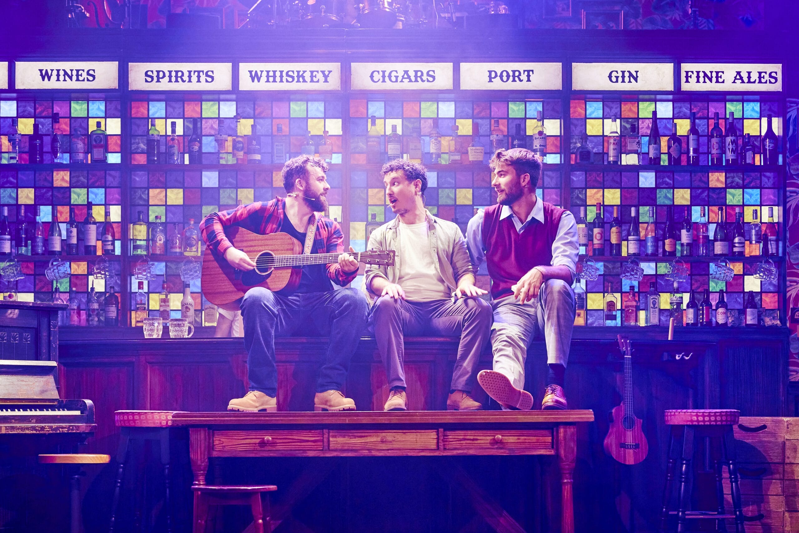 Three men sit on a wooden table in front of a colourful bar wall display; one strums a guitar while the others converse under signs for various drinks, reminiscent of The Choir of Man’s lively pub atmosphere.