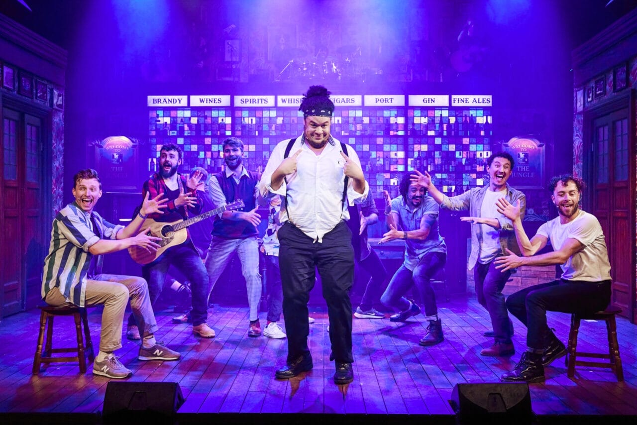 The Choir of Man performs a lively musical number on stage, with one man standing confidently in the center as others gesture animatedly toward him under colourful lighting.