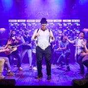 The Choir of Man performs a lively musical number on stage, with one man standing confidently in the center as others gesture animatedly toward him under colourful lighting.