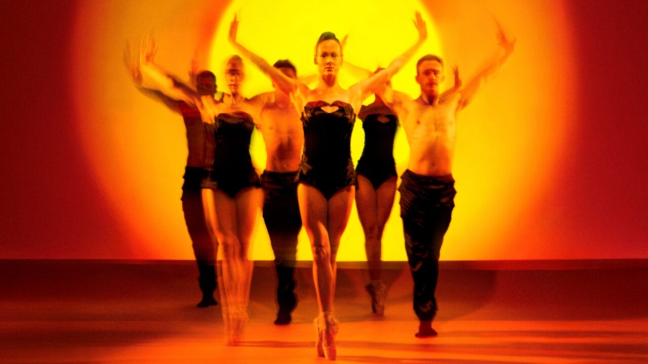 Five dancers in black costumes perform in front of a vivid orange and yellow backdrop, with blurred motion emphasizing dynamic movement.