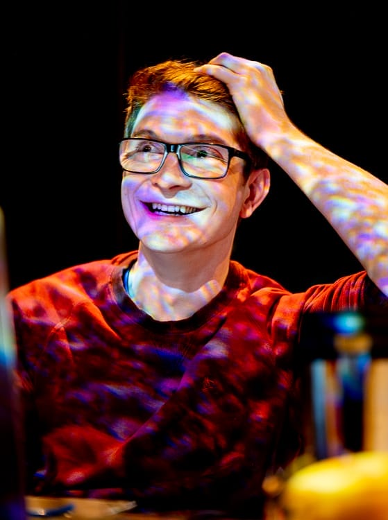 A smiling person wearing glasses and a red sweater sits with one hand on their head, illuminated by colourful, patterned lights.