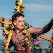 A man wearing a sheer floral shirt holds a large wooden cross adorned with flowers while standing waist-deep in water beneath a clear blue sky.