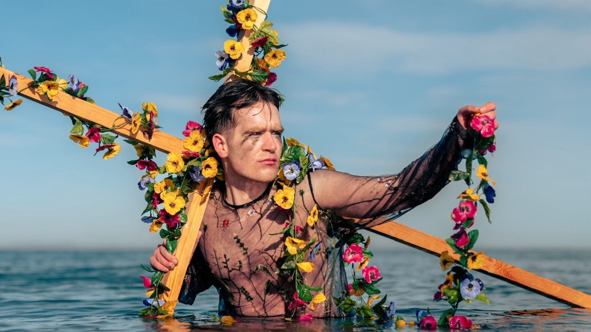 A man wearing a sheer floral shirt holds a large wooden cross adorned with flowers while standing waist-deep in water beneath a clear blue sky.