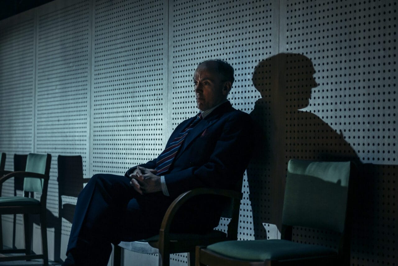 A man in a suit sits alone on a chair against a wall with circular perforations, casting a shadow under moody lighting—a scene reminiscent of The Spy Who Came in from the Cold.
