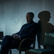 A man in a suit sits alone on a chair against a wall with circular perforations, casting a shadow under moody lighting—a scene reminiscent of The Spy Who Came in from the Cold.