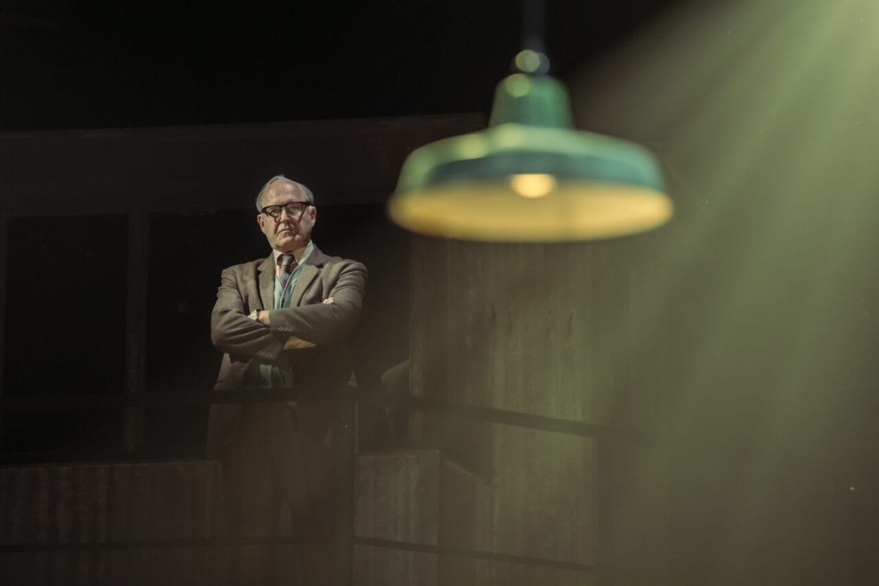 An older man in a suit and glasses stands with arms crossed under dim lighting, evoking the atmosphere of a John le Carré spy novel, with a green hanging lamp in the foreground and light rays streaming down.