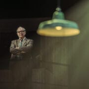 An older man in a suit and glasses stands with arms crossed under dim lighting, evoking the atmosphere of a John le Carré spy novel, with a green hanging lamp in the foreground and light rays streaming down.