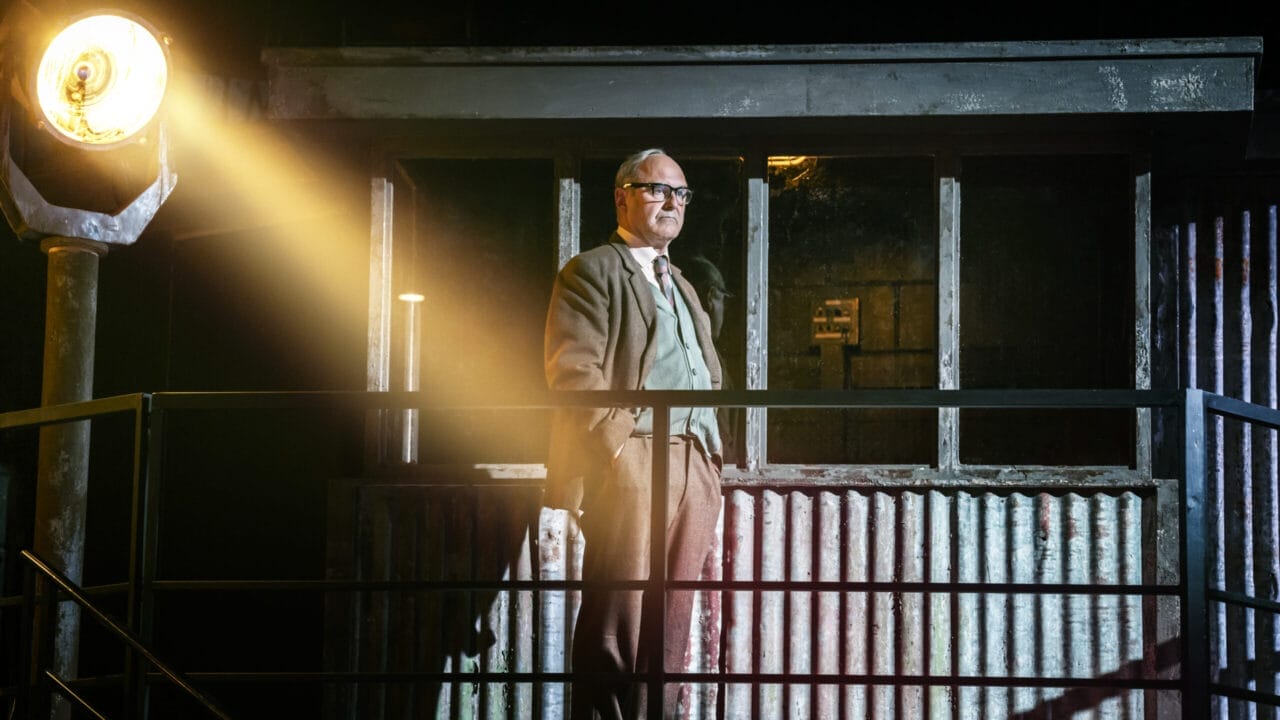 A man in a suit and glasses stands on a metal balcony, illuminated by a bright spotlight, evoking the tense atmosphere of The Spy Who Came in from the Cold, with a corrugated metal structure and windows behind him.