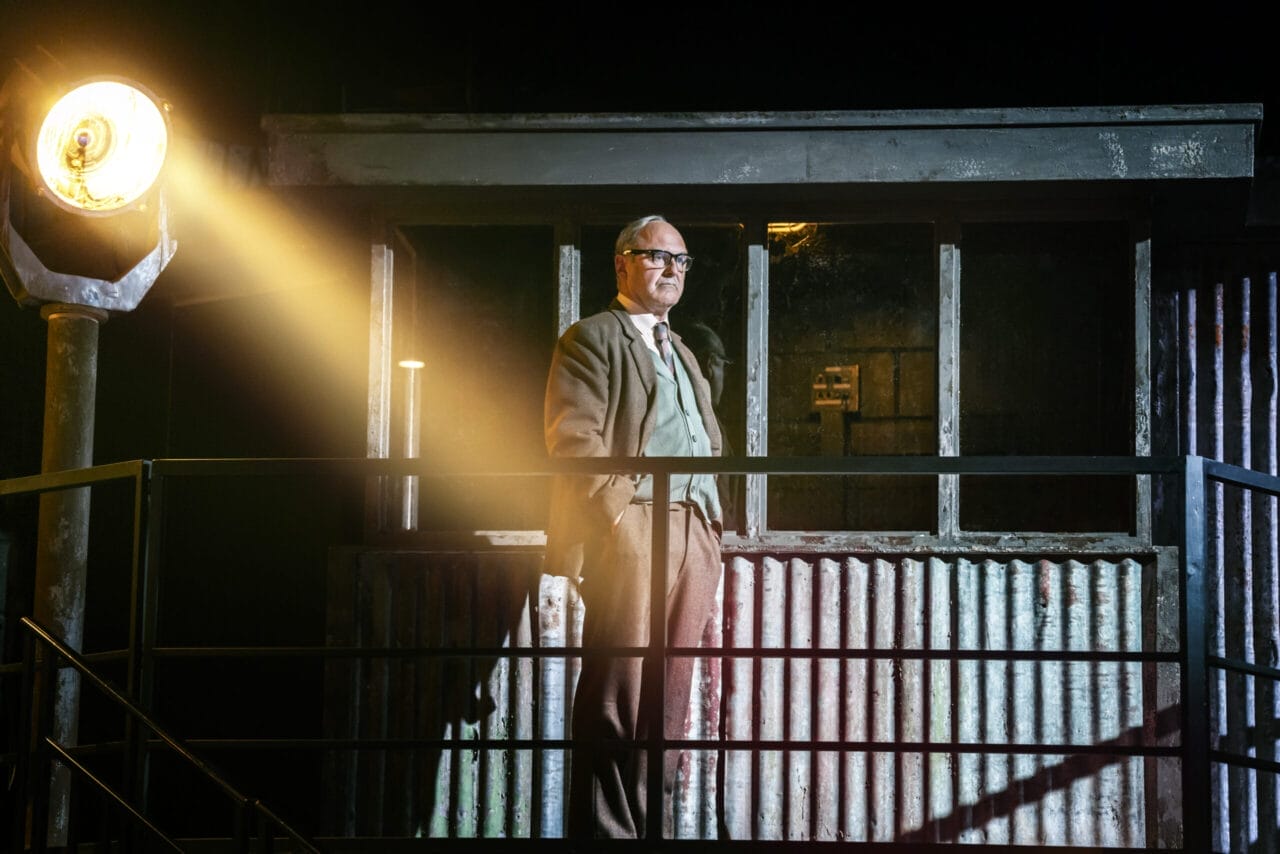A man in a suit and glasses stands on a metal balcony, illuminated by a bright spotlight, evoking the tense atmosphere of The Spy Who Came in from the Cold, with a corrugated metal structure and windows behind him.