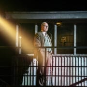 A man in a suit and glasses stands on a metal balcony, illuminated by a bright spotlight, evoking the tense atmosphere of The Spy Who Came in from the Cold, with a corrugated metal structure and windows behind him.