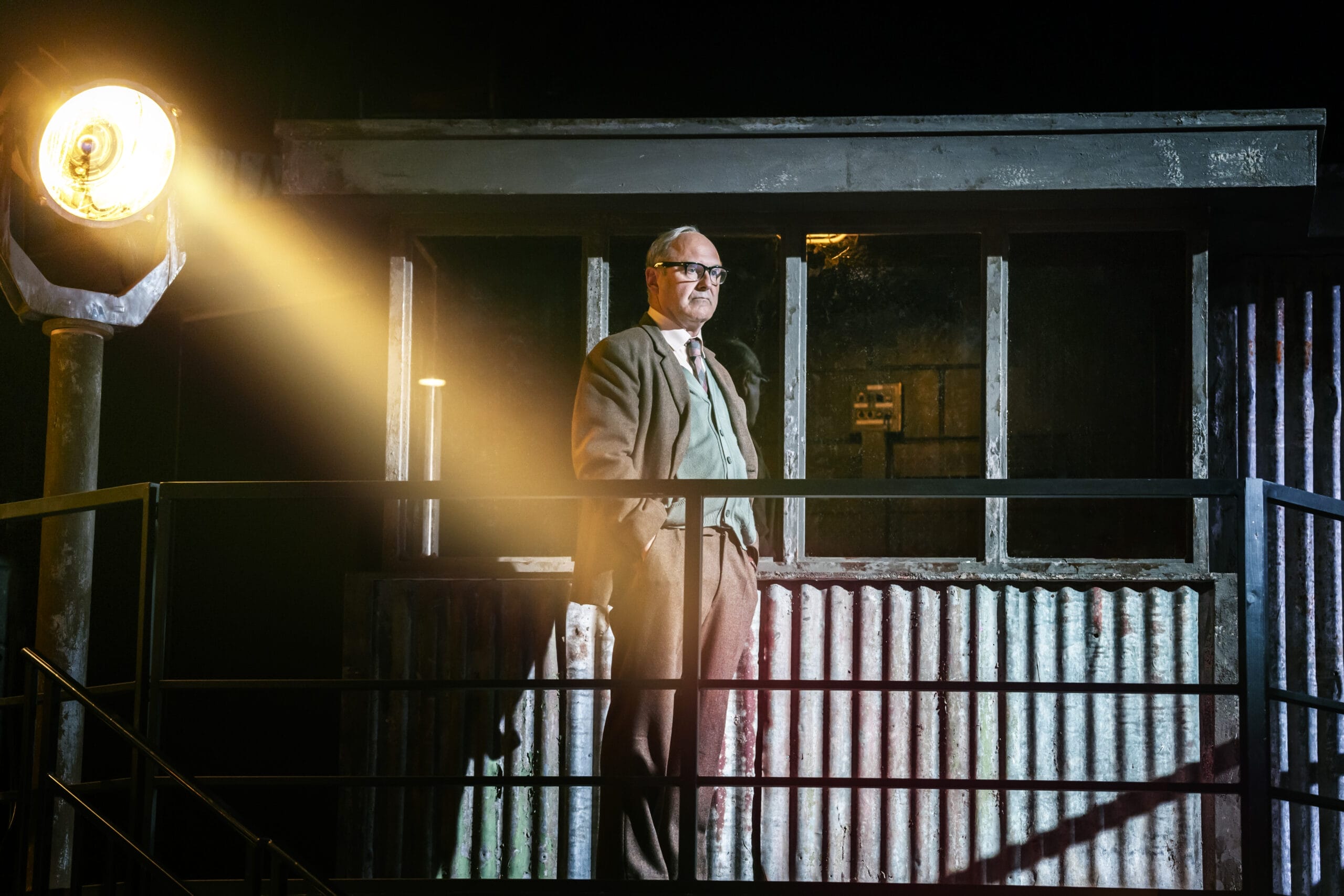 A man in a suit and glasses stands on a metal balcony, illuminated by a bright spotlight, evoking the tense atmosphere of The Spy Who Came in from the Cold, with a corrugated metal structure and windows behind him.