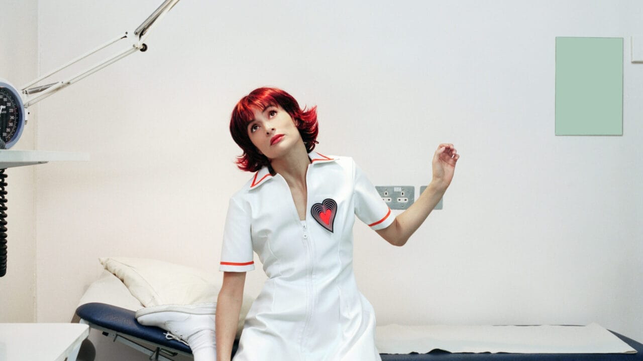 A woman with short red hair in a white dress with a heart patch sits on a medical examination table in a brightly lit, minimal clinic room.