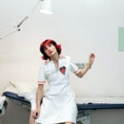 A woman with short red hair in a white dress with a heart patch sits on a medical examination table in a brightly lit, minimal clinic room.
