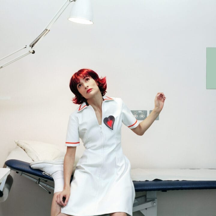 A woman with short red hair in a white dress with a heart patch sits on a medical examination table in a brightly lit, minimal clinic room.