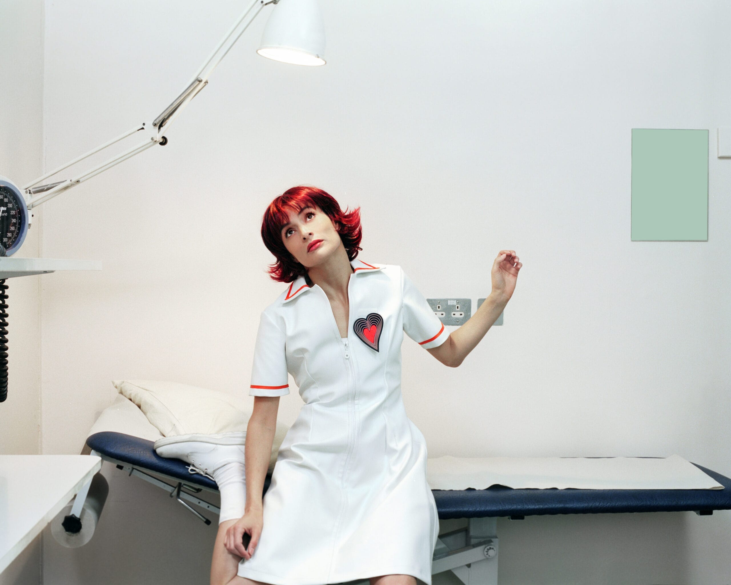 A woman with short red hair in a white dress with a heart patch sits on a medical examination table in a brightly lit, minimal clinic room.