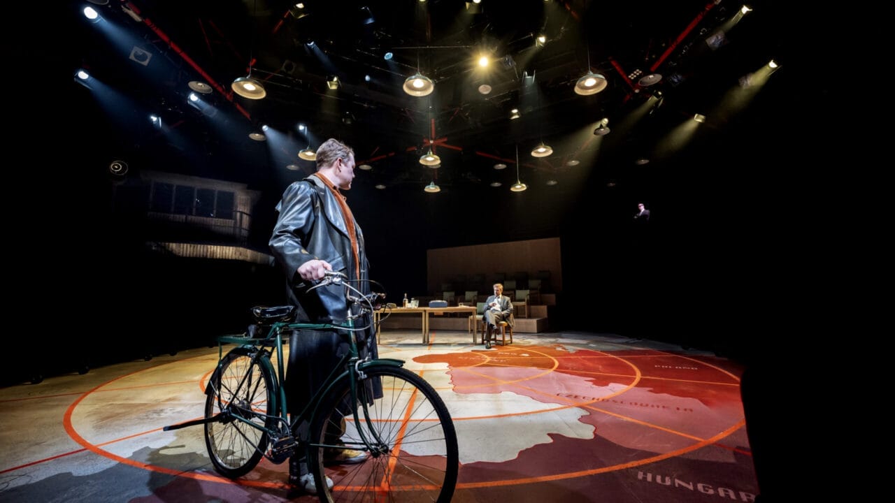 A person with a bicycle stands on a stage under dramatic lighting, evoking the suspense of a spy novel, while another person sits at a desk in the background.