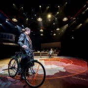 A person with a bicycle stands on a stage under dramatic lighting, evoking the suspense of a spy novel, while another person sits at a desk in the background.