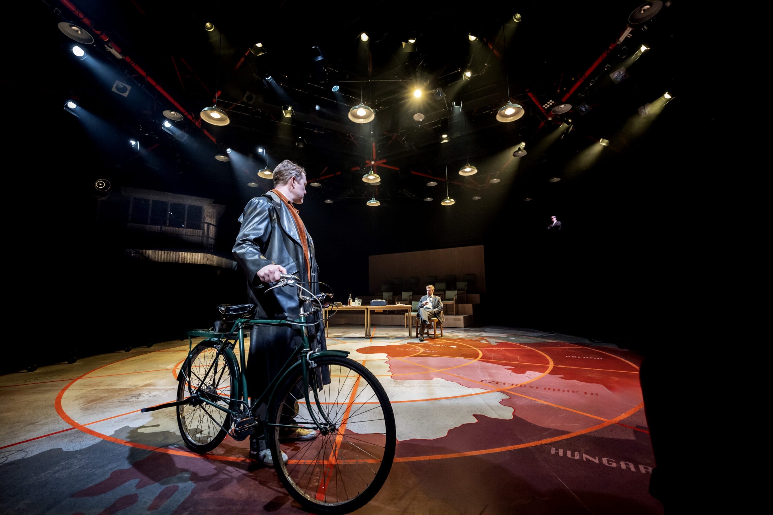 A person with a bicycle stands on a stage under dramatic lighting, evoking the suspense of a spy novel, while another person sits at a desk in the background.