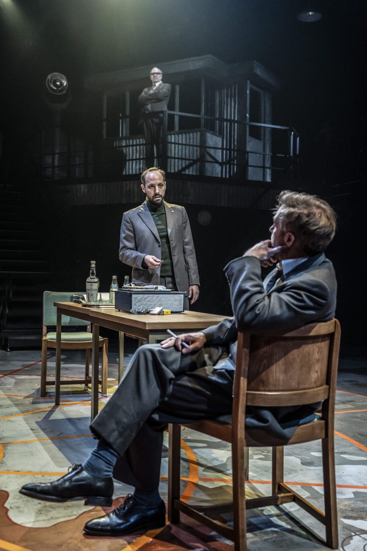 Three men in suits engage in a tense conversation in a dimly lit room—an atmosphere reminiscent of The Spy Who Came in from the Cold—one sits at a desk, another stands, while the third observes silently from a balcony above.