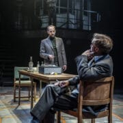 Three men in suits engage in a tense conversation in a dimly lit room—an atmosphere reminiscent of The Spy Who Came in from the Cold—one sits at a desk, another stands, while the third observes silently from a balcony above.