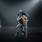 A group of Rambert dancers perform on a dark stage; one person lifts another, while others pose and move gracefully in the background under spotlights.