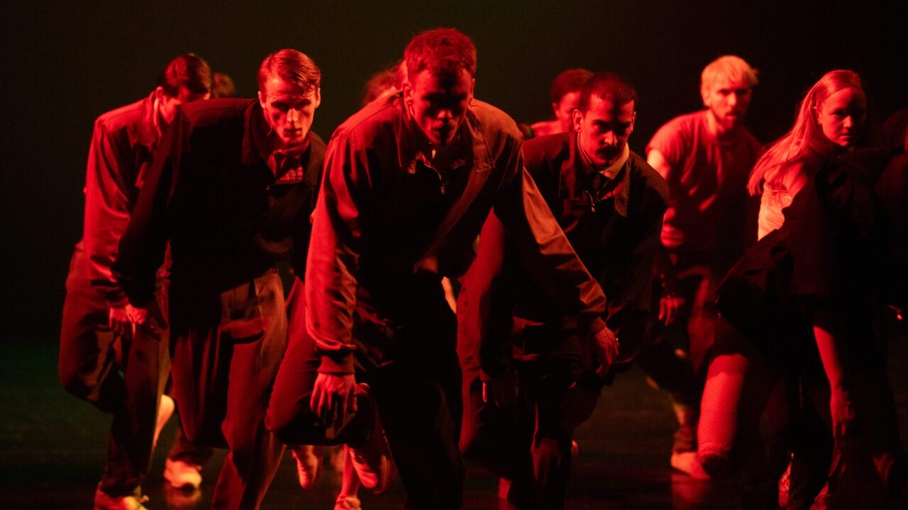 A group of Rambert dancers in tracksuits perform on stage under red lighting, moving in a low, intense formation.