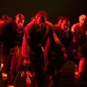 A group of Rambert dancers in tracksuits perform on stage under red lighting, moving in a low, intense formation.
