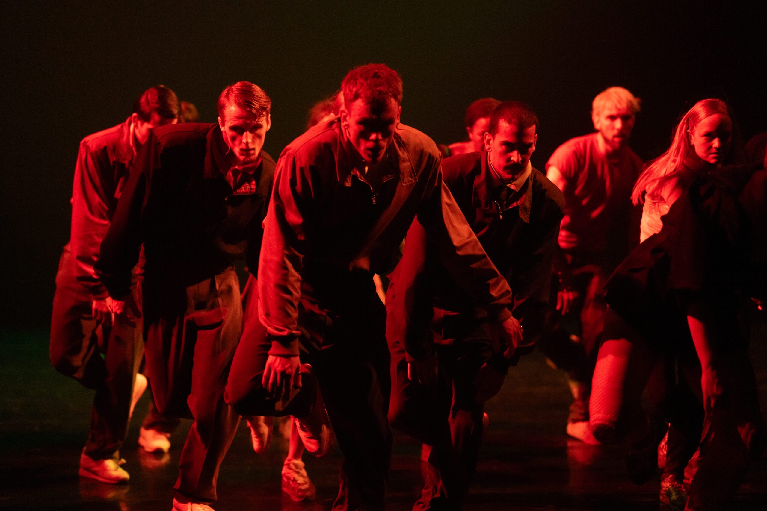 A group of Rambert dancers in tracksuits perform on stage under red lighting, moving in a low, intense formation.