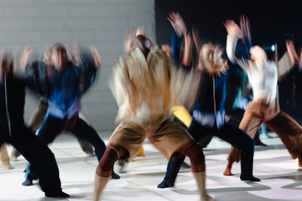 A group of people in motion, blurred as they perform energetic Rambert dance movements on a light-colored floor.