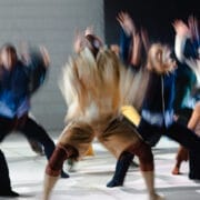 A group of people in motion, blurred as they perform energetic Rambert dance movements on a light-colored floor.