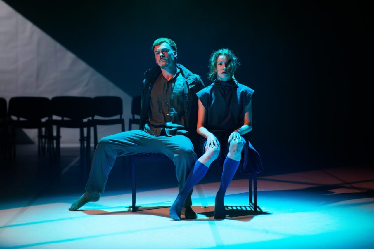 Two actors sit on a bench under dramatic stage lighting, facing forward with serious expressions; empty chairs are visible in the background, evoking the intensity of a Rambert performance.