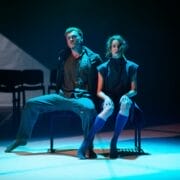 Two actors sit on a bench under dramatic stage lighting, facing forward with serious expressions; empty chairs are visible in the background, evoking the intensity of a Rambert performance.