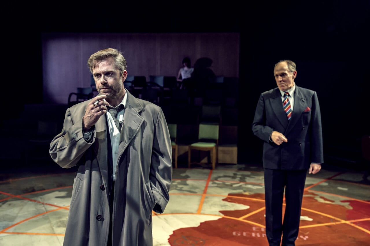 Two men in suits stand on a stage with a large map on the floor, evoking a tense scene from a John le Carré spy novel; one smokes while the other looks on. Audience seating and a person are visible in the background.