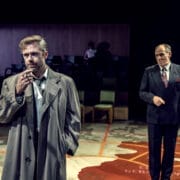 Two men in suits stand on a stage with a large map on the floor, evoking a tense scene from a John le Carré spy novel; one smokes while the other looks on. Audience seating and a person are visible in the background.
